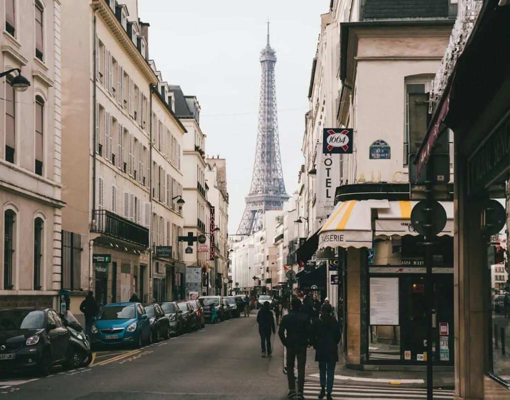 A bustling Parisian street scene with pedestrians walking down the sidewalk, flanked by traditional buildings and a small hotel. In the distance, the Eiffel Tower looms large, capturing the essence of Paris in a single frame. The street life and iconic architecture embody the charm and allure that awaits visitors on a day trip from London to Paris, as featured in the blog "Insight Trends."